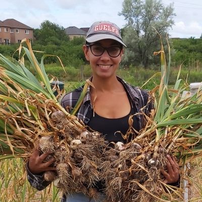 Rav Singh stands smiling in her field holding a bundle of freshly harvested garlic in each hand