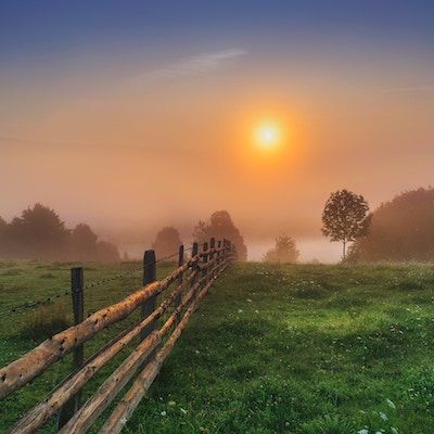 an orange sun rises above a lush green pasture bordered by a split rail fence